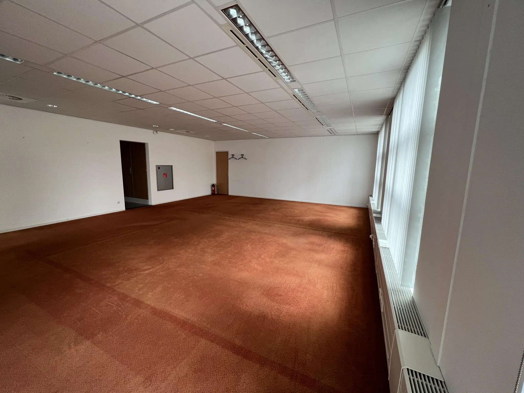 Leeg kantoorruimte met roodbruine vloerbedekking aan de Rivium 2e Straat, met ramen en verticale jaloezieën aan de rechterkant.